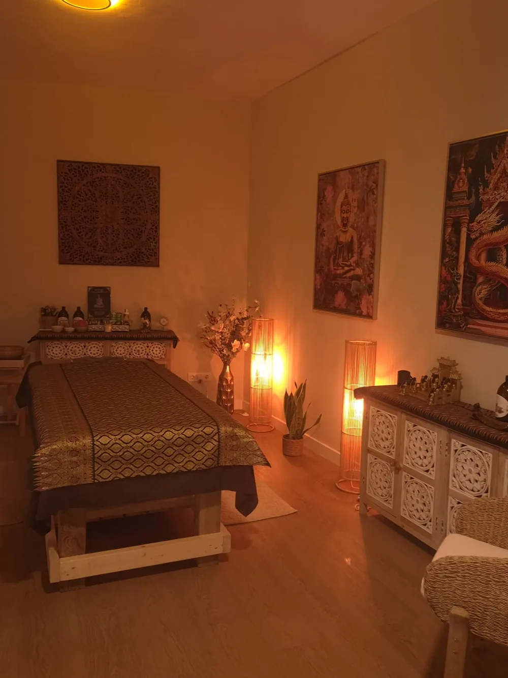Sala de massatge amb llitera de seda daurada, llums de llum càlida, quadres tailandesos i butaca de vímet